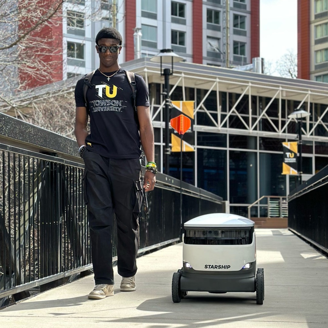 Towson University student walking outside next to a Starship food delivery robot.